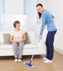 caregiver cleaning the floor with a mop while a senior woman sitting on the sofa